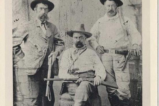 3 men face the camera.  All three are wearing cowboy hats and western garb.