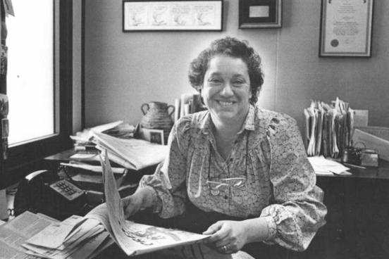 a woman holding a newspaper, sitting in an office.  A typewriter and phone are behind her.